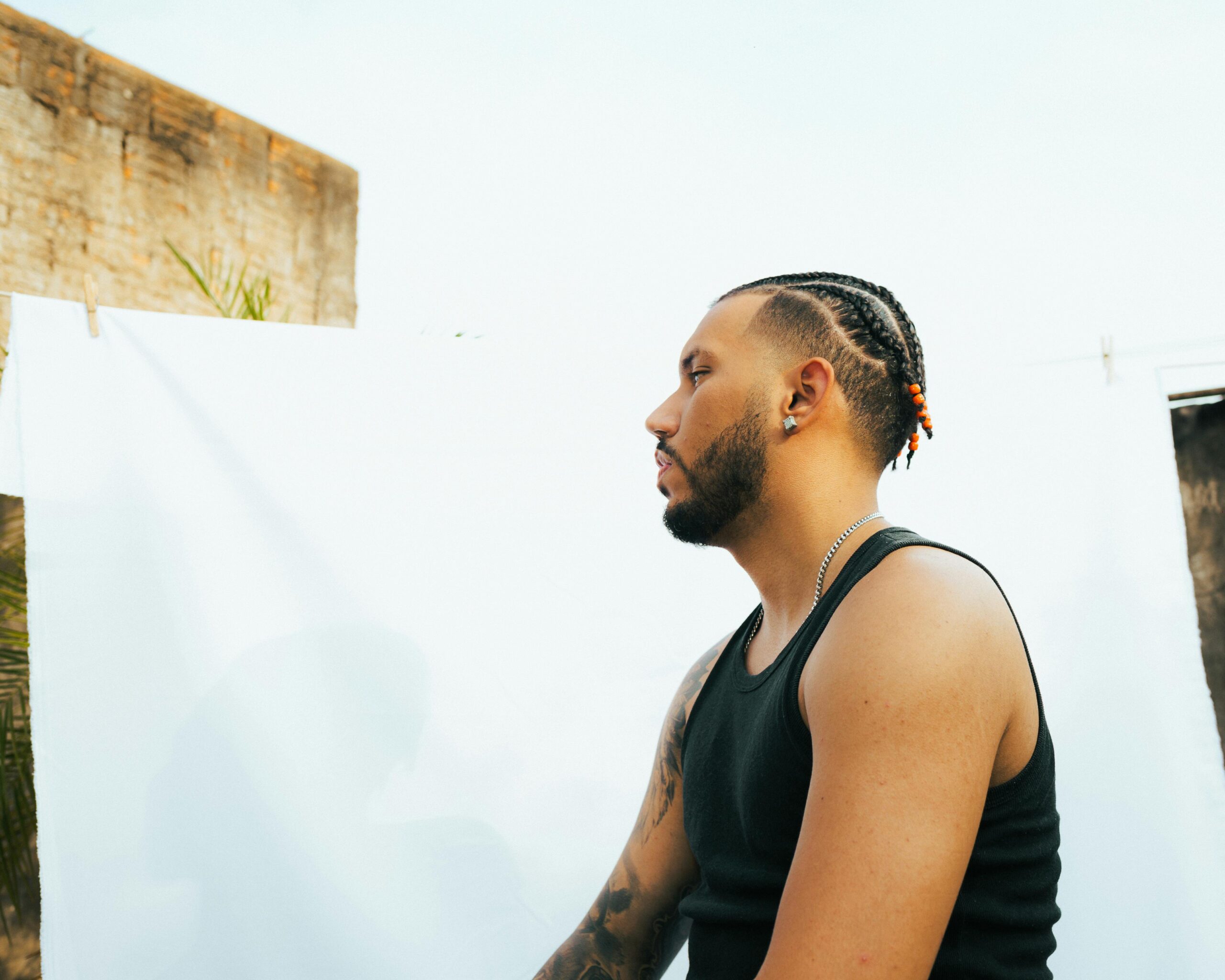 Profile portrait of a tattooed man with braids outdoors against a white backdrop.