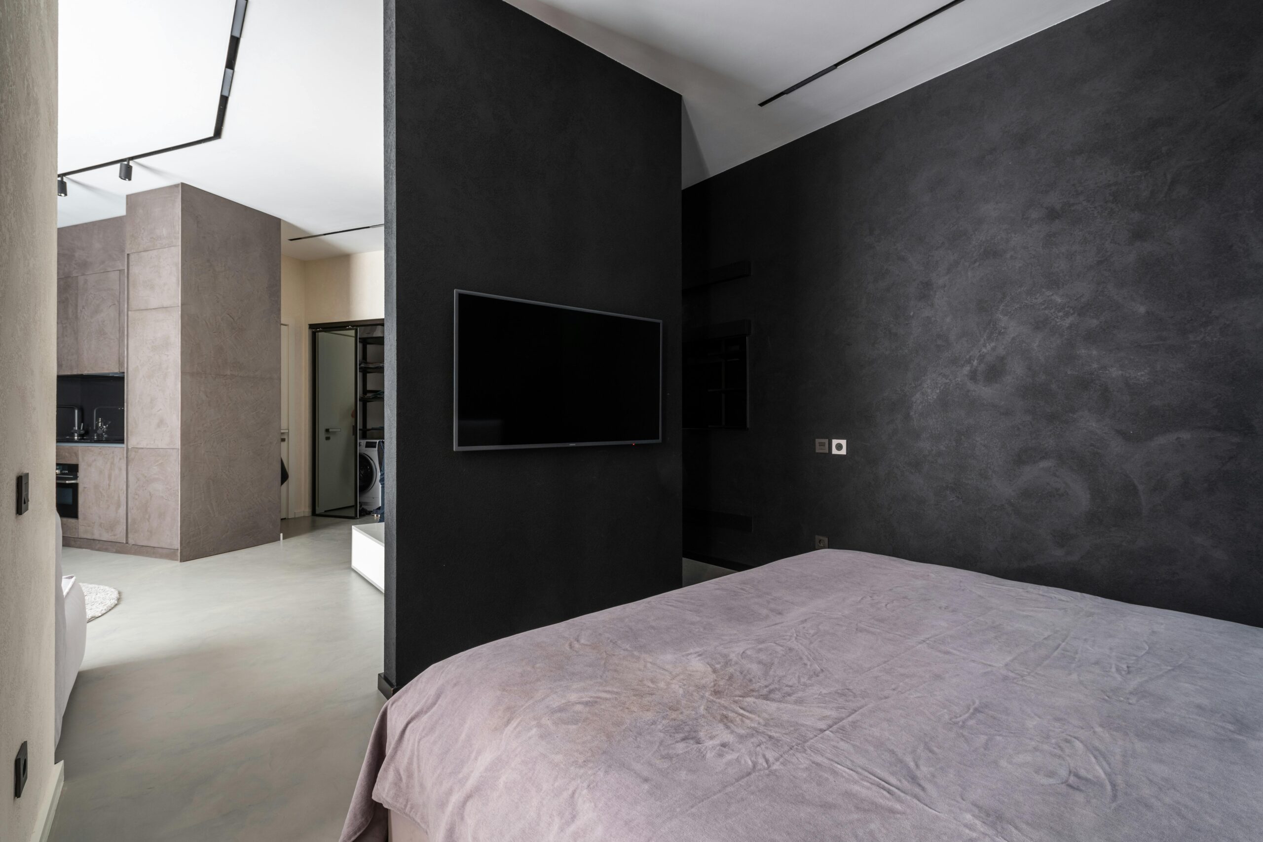 Sleek contemporary bedroom with a black TV, gray walls, and open kitchen view.